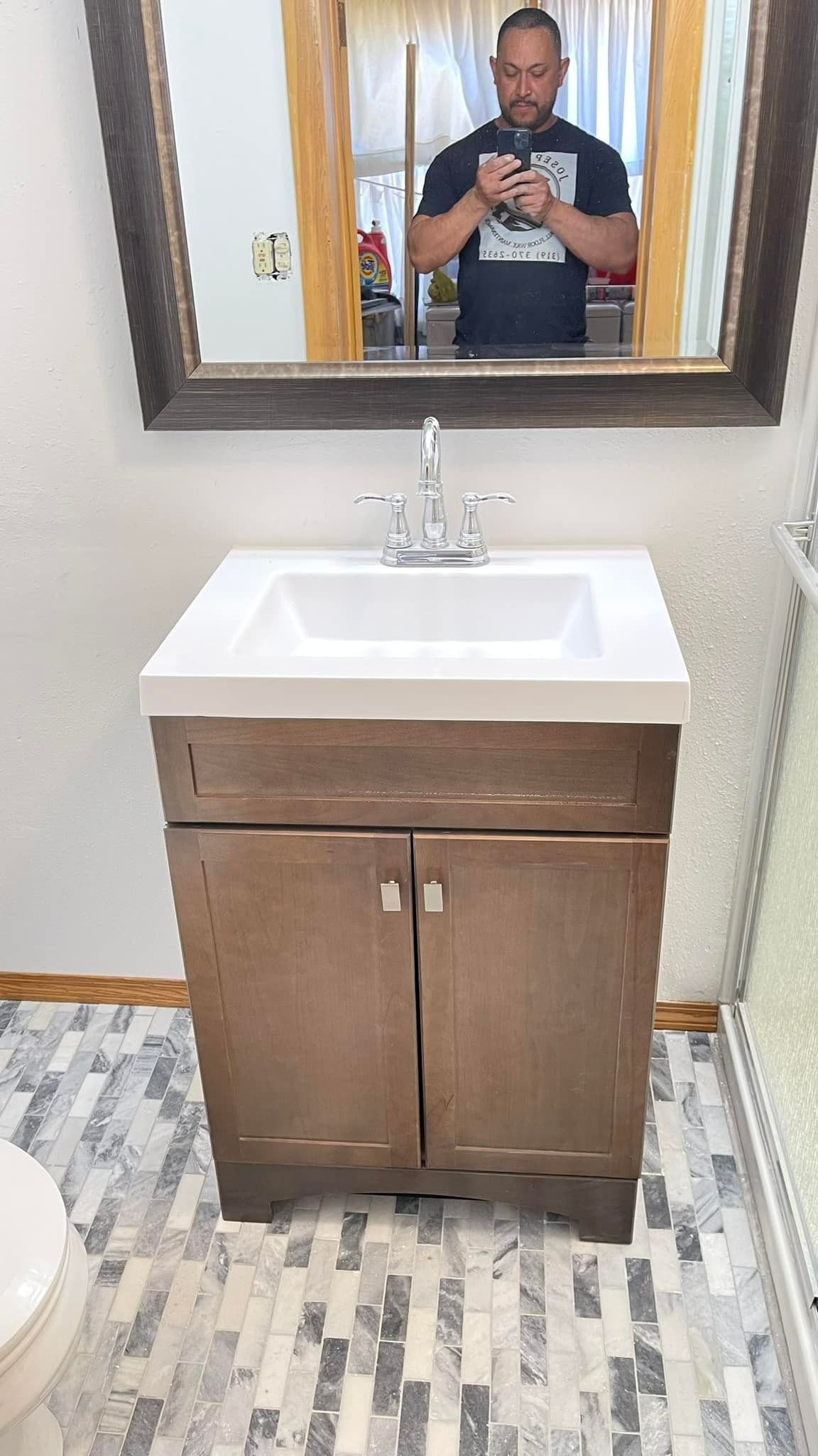 A person stands in a bathroom in front of a mirror above a wooden vanity with a white rectangular sink.