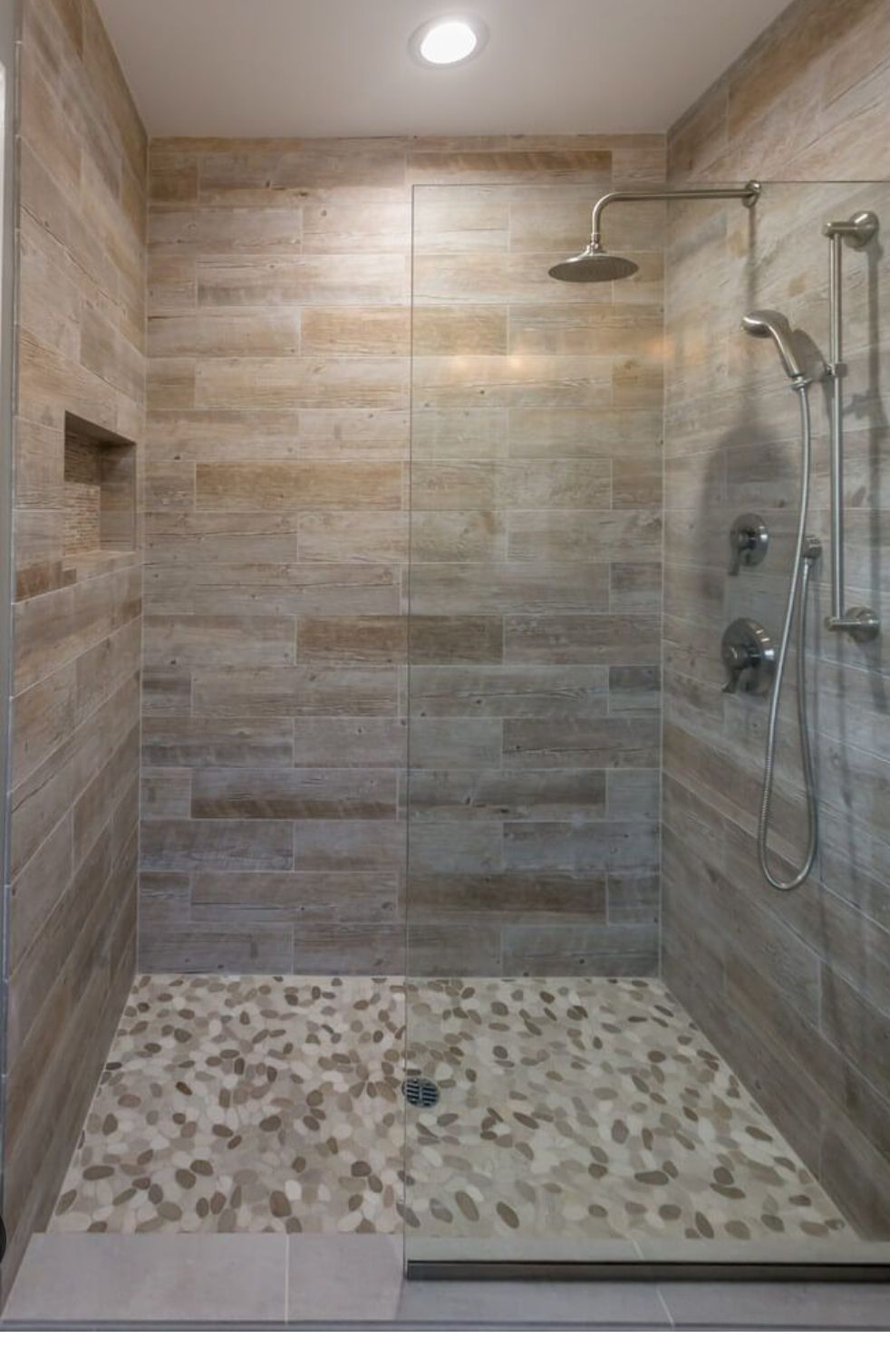 A modern walk-in shower with horizontal beige stone tile walls, a pebble-stone floor, glass panel, and chrome fixtures.