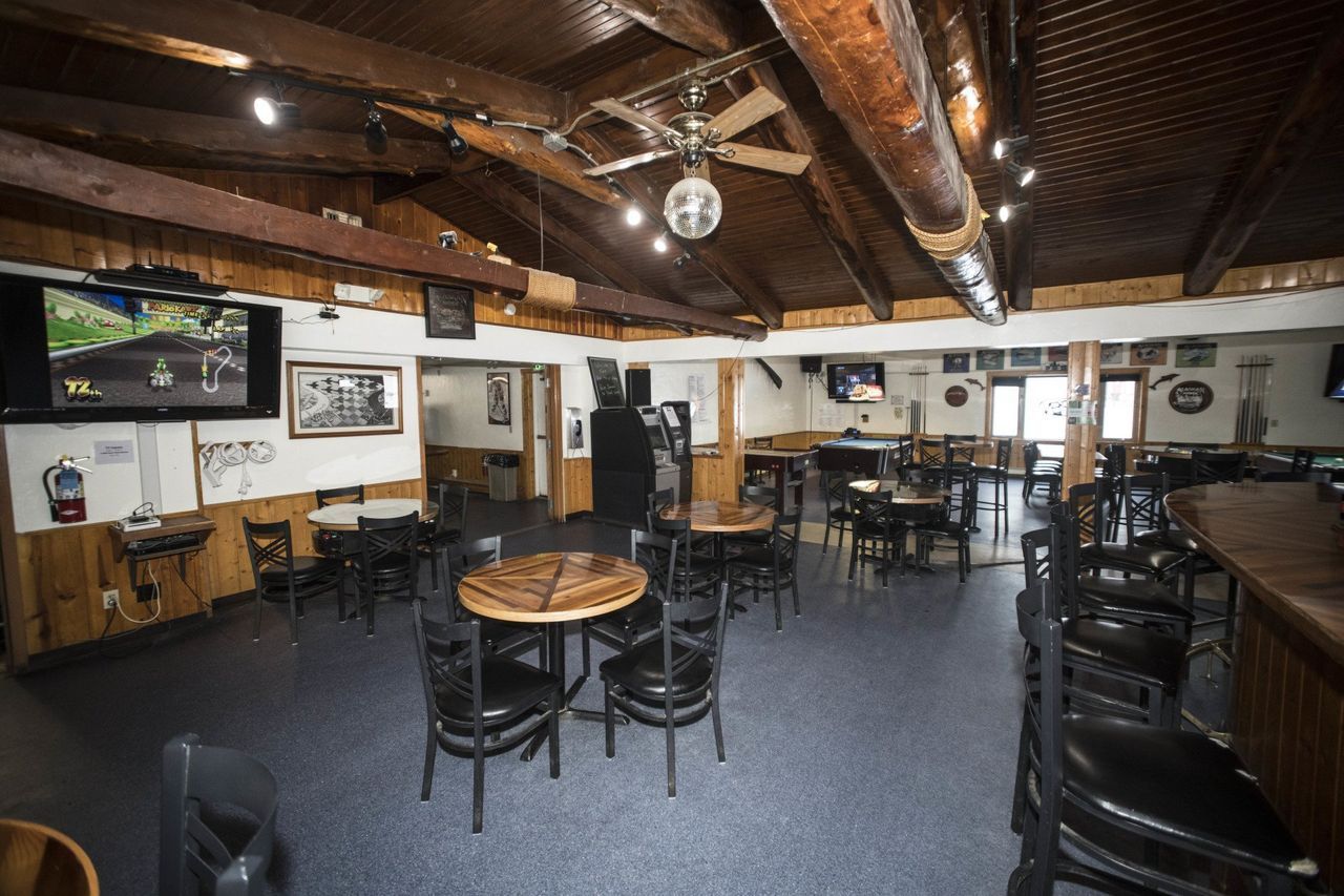Pub | Healy, AK | Denali Totem Inn Interior view of a dimly lit bar with round tables, chairs, and a long counter. TVs and a disco ball are visible.