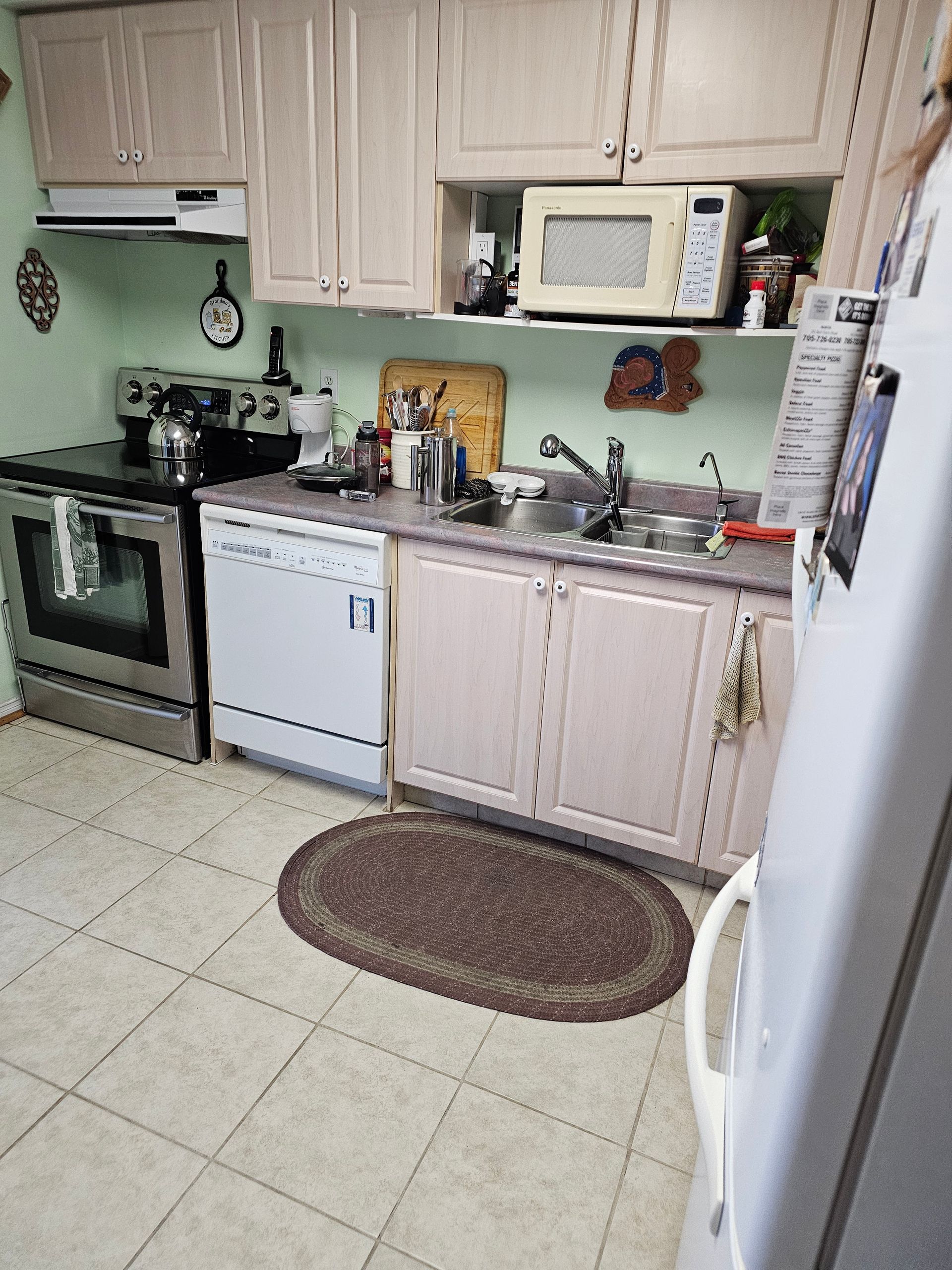 Small kitchen with beige cabinets, white appliances, sink, stove, microwave, and oval rug on tiled floor