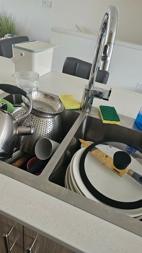 Kitchen sink piled with dishes and utensils under a chrome faucet, with plates and cookware scattered around.