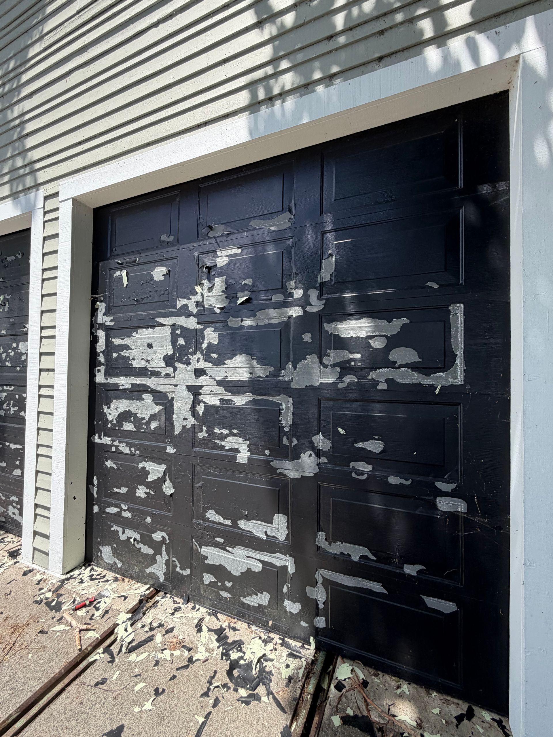 Black garage door with peeling paint on a house exterior, beside a gravel walkway.