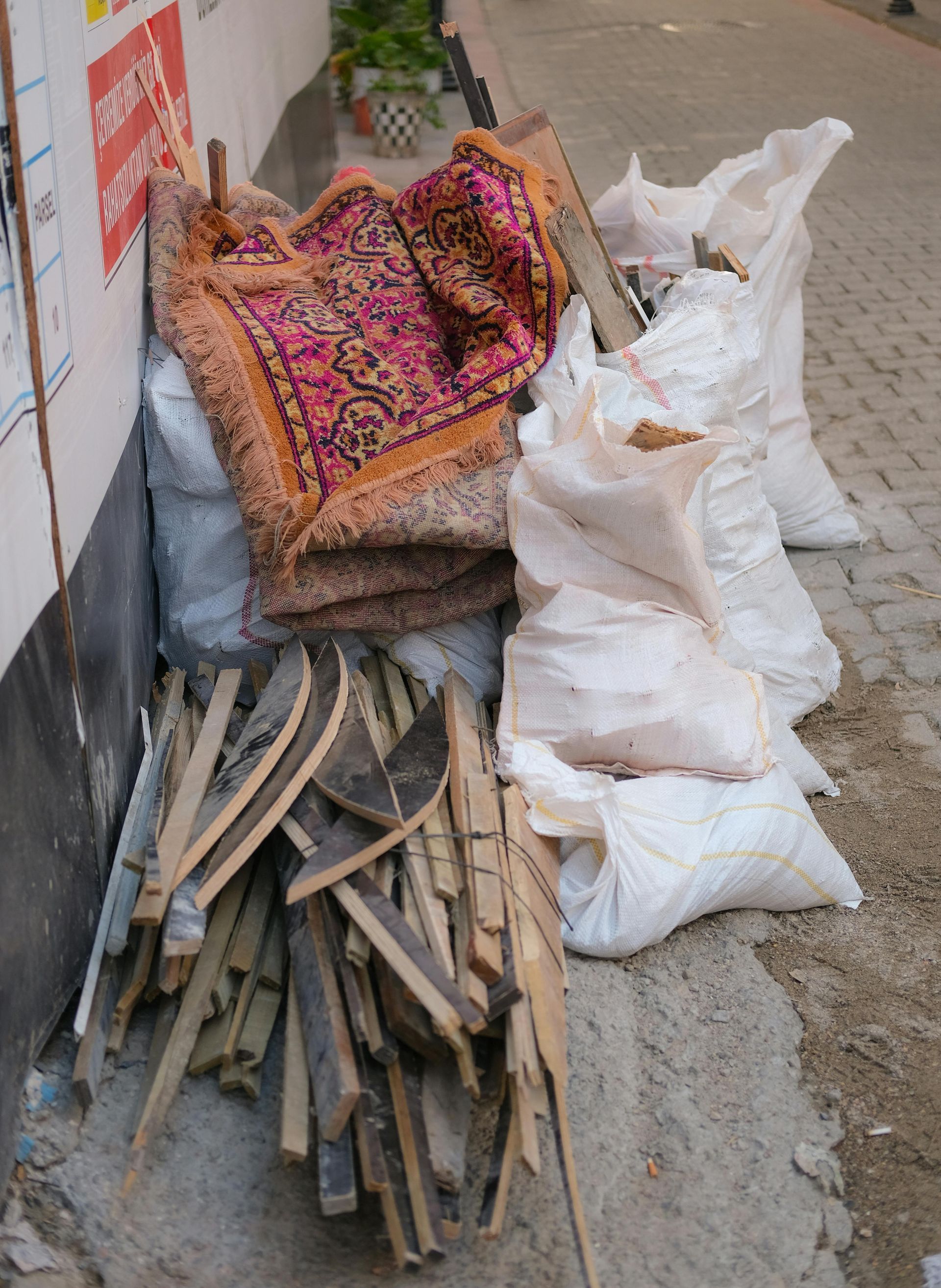Piles of folded rugs, sacks, and wooden sticks stacked along a sidewalk beside a wall.