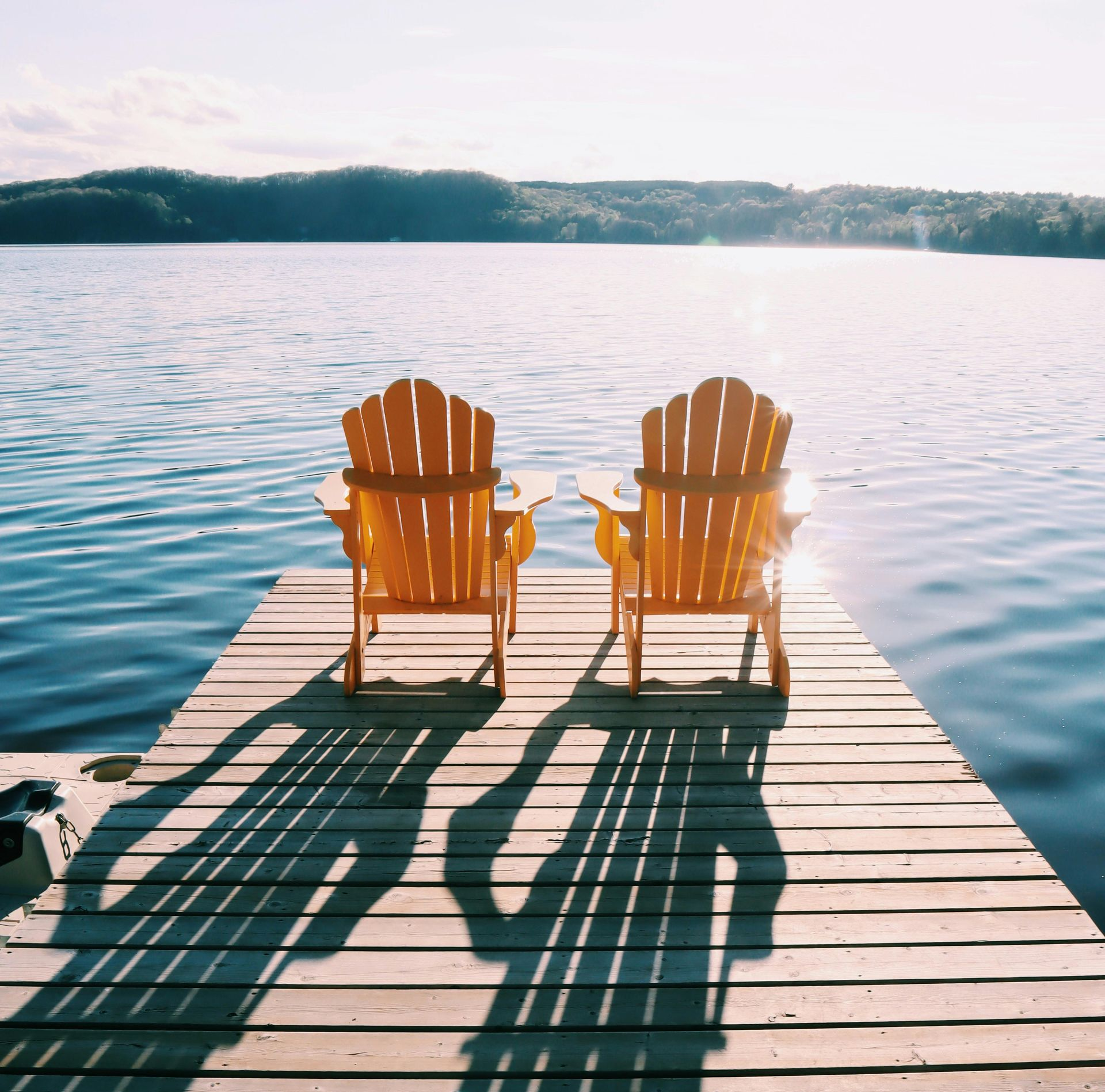 Two wooden chairs on a dock facing a calm lake at sunrise, with long shadows stretching behind them.