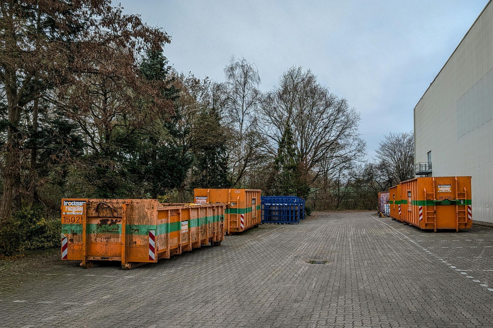 Gravel lot with rows of orange construction dumpsters and blue bins beside a large white building