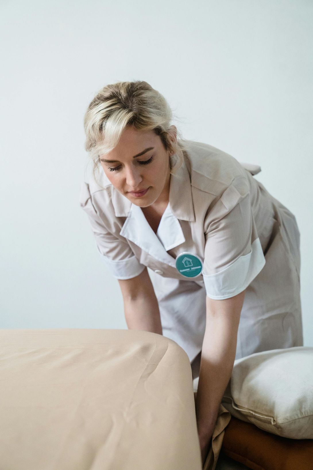Nurse in white uniform adjusting a bed sheet beside a beige bed in a bright room