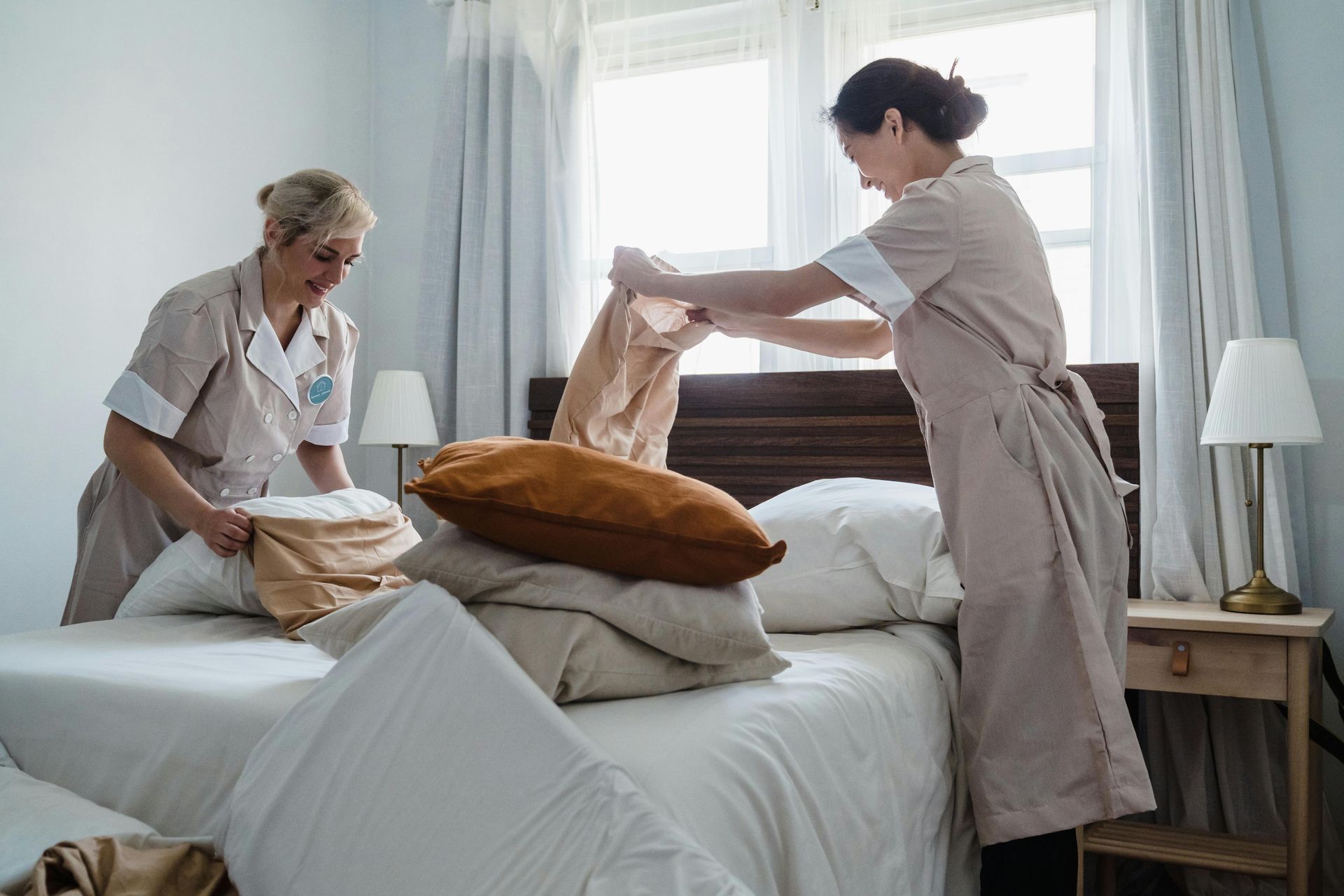 Two people making a bed in a bright bedroom, arranging pillows and bedding.