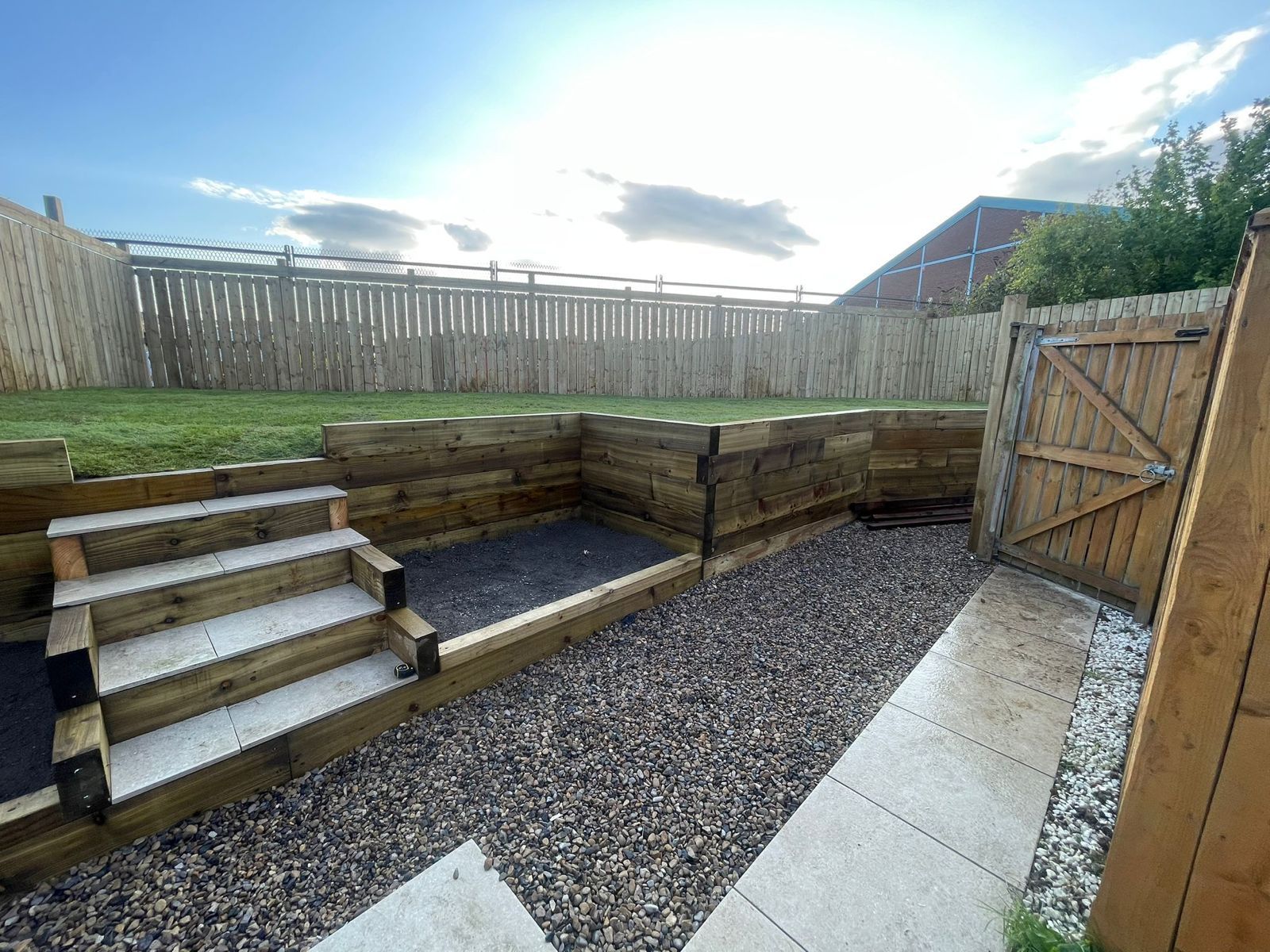Backyard with gravel seating area, wooden steps, retaining wall, and fenced gate under a bright sky