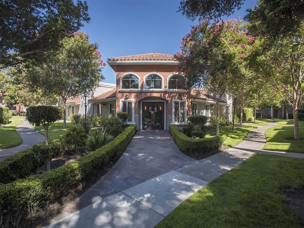 Entrance to a sunlit apartment community with curved walkways, lush landscaping, and a pink stucco building.
