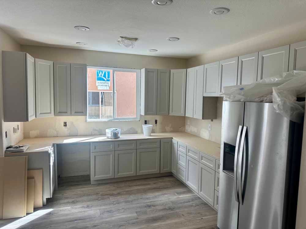 Empty kitchen with gray cabinets, stainless refrigerator, and a sink beneath a window under construction.