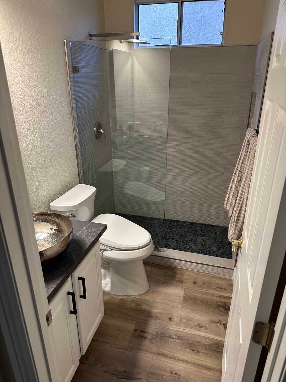 Small bathroom with toilet, sink, glass shower, wood floor, and a window above the shower.