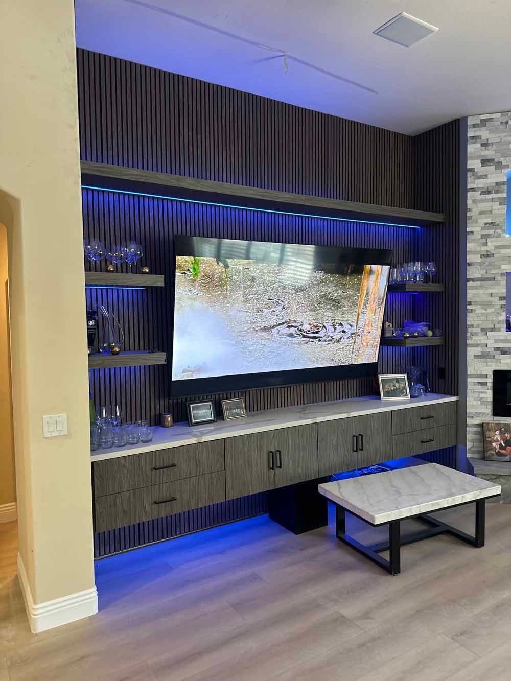Built-in living room media wall with large TV, blue accent lighting, shelves, and a small coffee table
