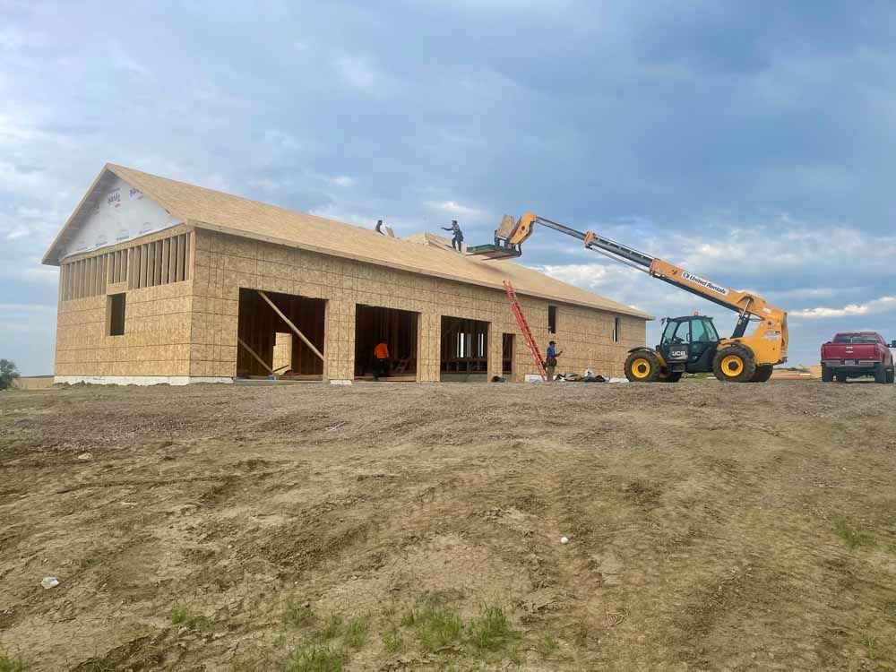 Construction site with a house frame, telehandler lifting materials, and a worker on the roof under a cloudy sky