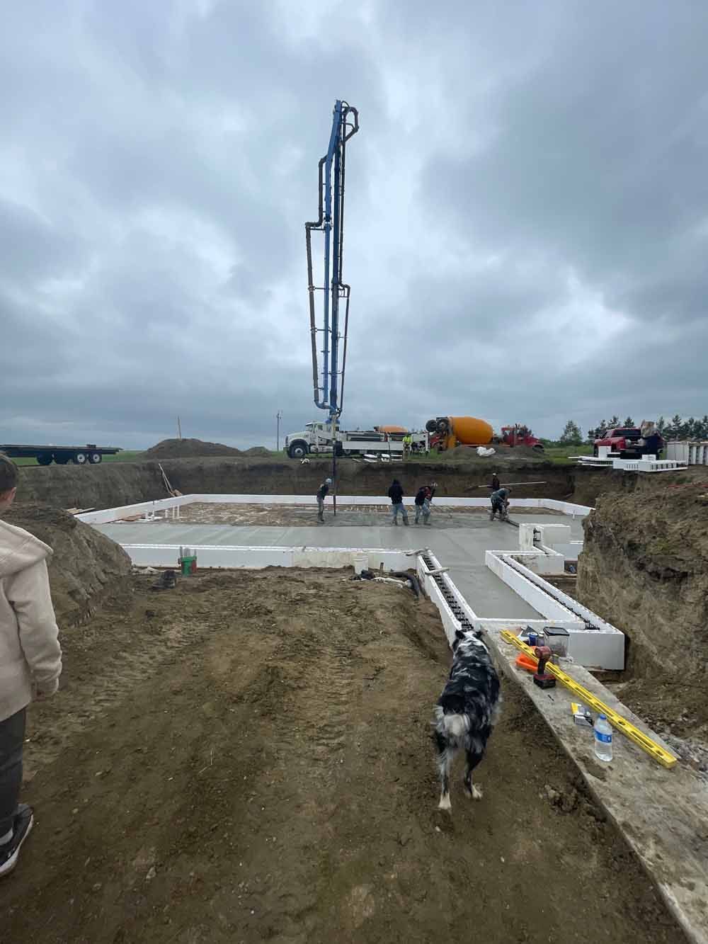Construction site with foundation pit, concrete forms, workers, and a tall pump truck under cloudy sky