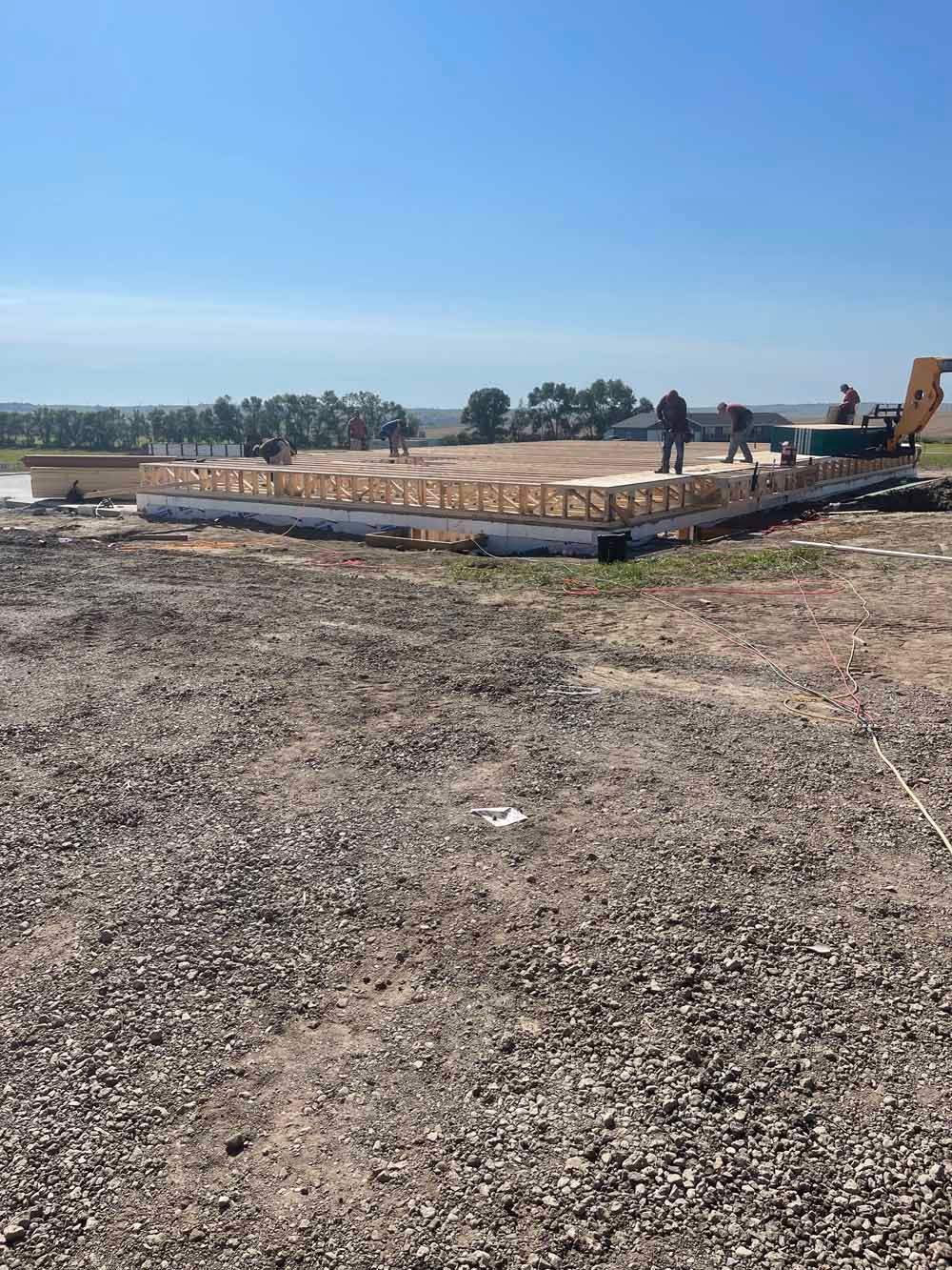 Construction site with excavator, workers, and concrete foundation on a gravel lot under a clear blue sky