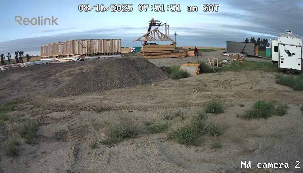 Construction site with dirt mounds, excavator, stacked materials, and a white truck near the water