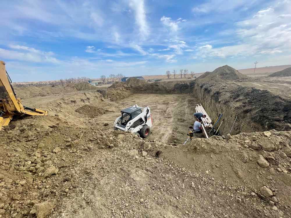Two people work beside a buried white vehicle in a muddy trench, with an excavator at left under a cloudy sky
