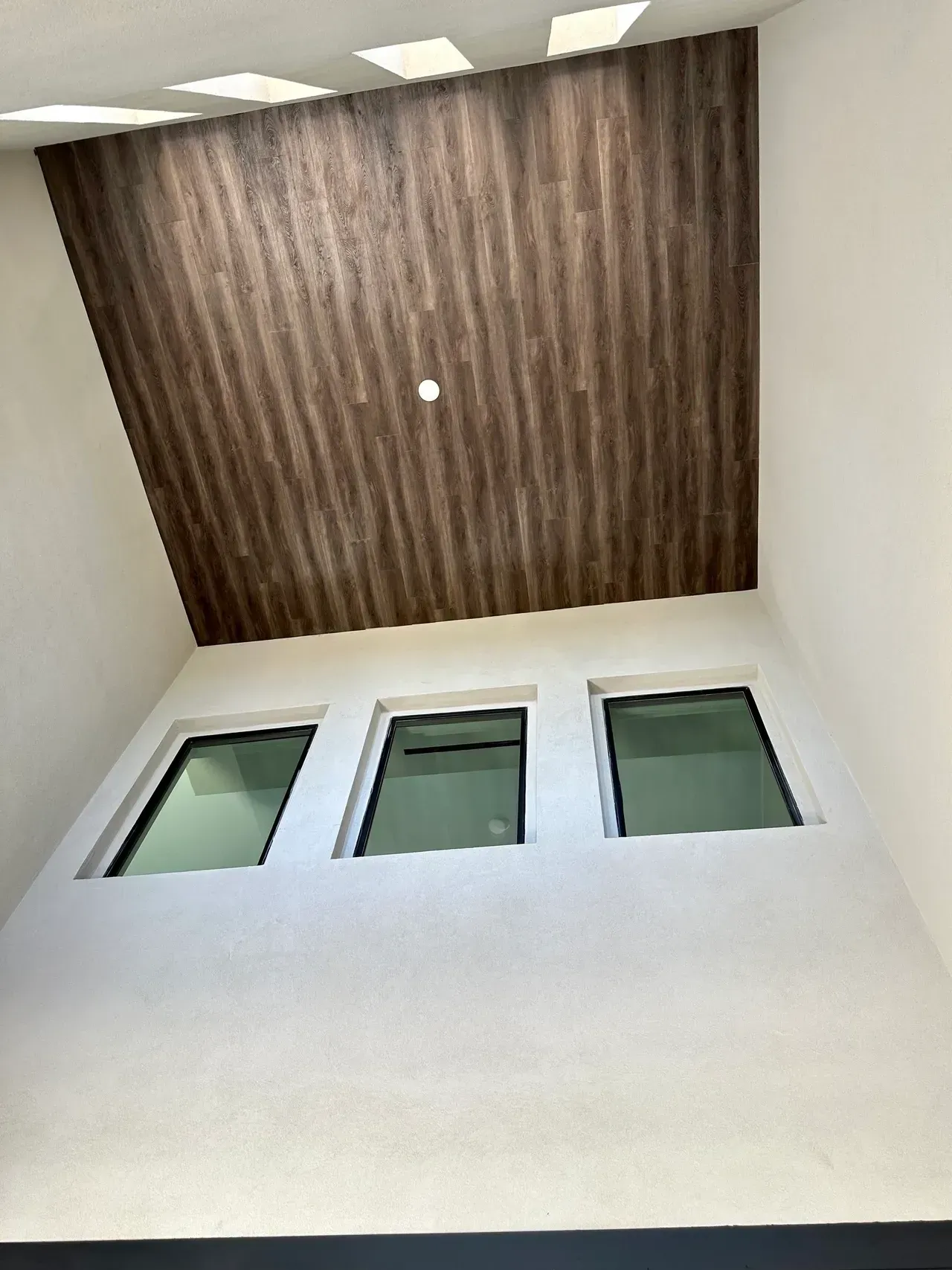 Upward view of a tall white interior wall with three windows and a wood-paneled ceiling above