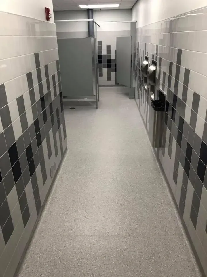 Narrow restroom hallway with gray tiled walls, stalls on the left, and a sink area at the end.