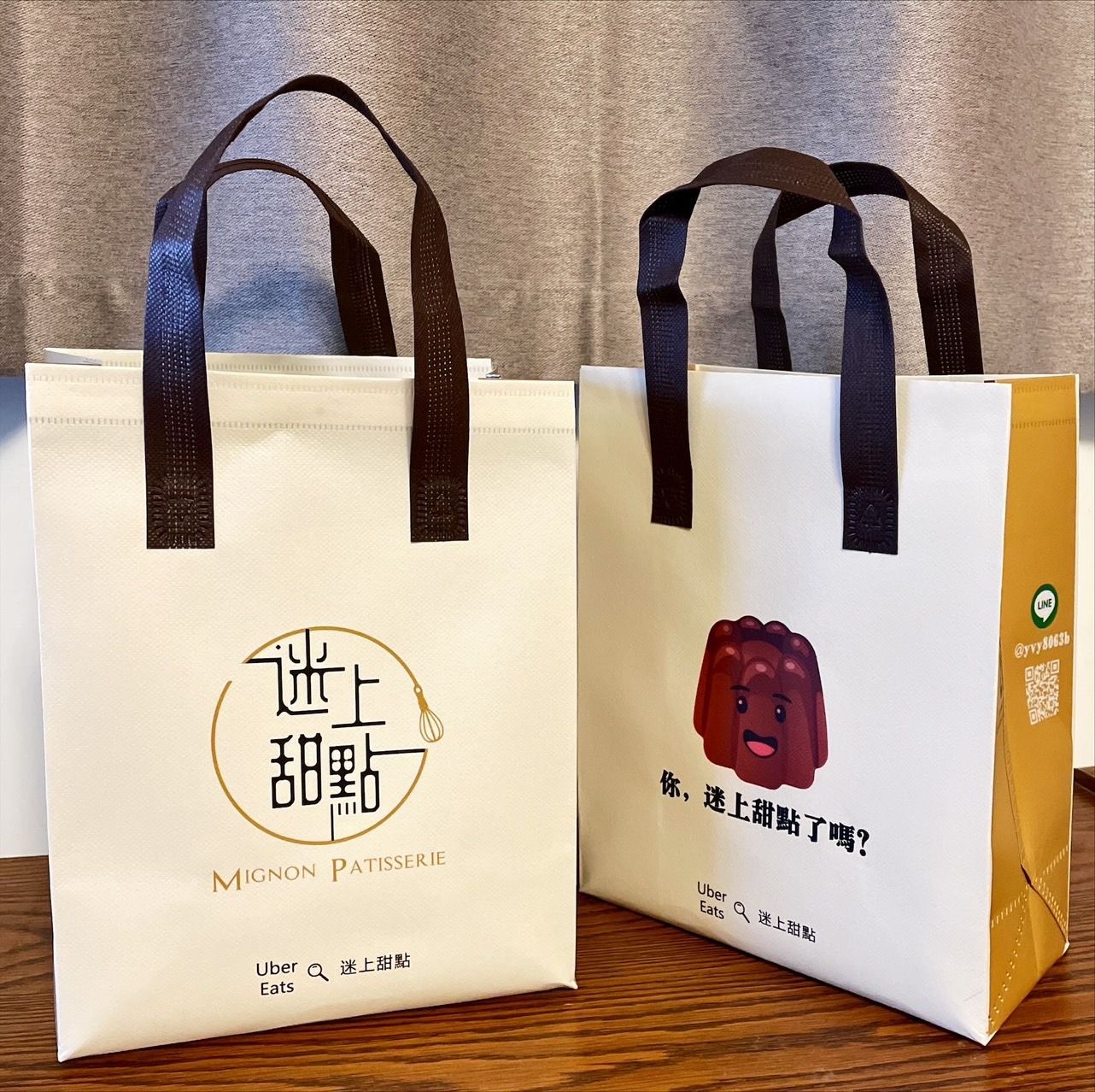 木桌上擺放著兩個米色禮品袋,袋子配有深棕色提手,其中一個印有中文,側邊為黃色。