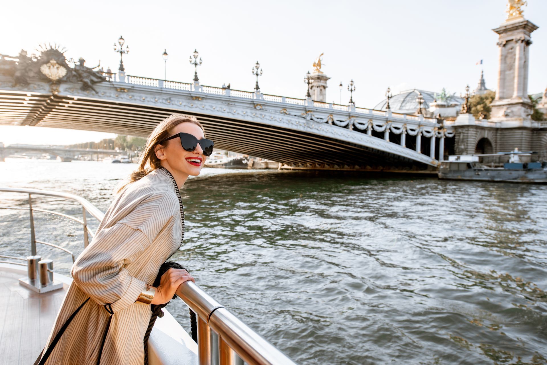 Woman in sunglasses leaning on a riverside railing with a bridge and water behind her