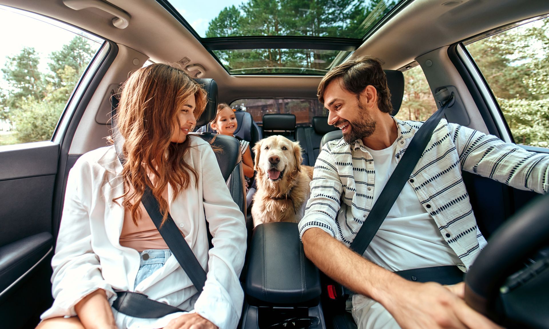 Two adults and a dog sitting in a car with a sunroof, smiling at each other