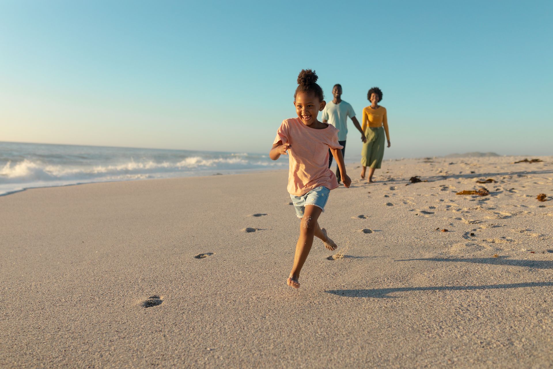 Children running along a sunny beach near the ocean, with footprints in the sand.