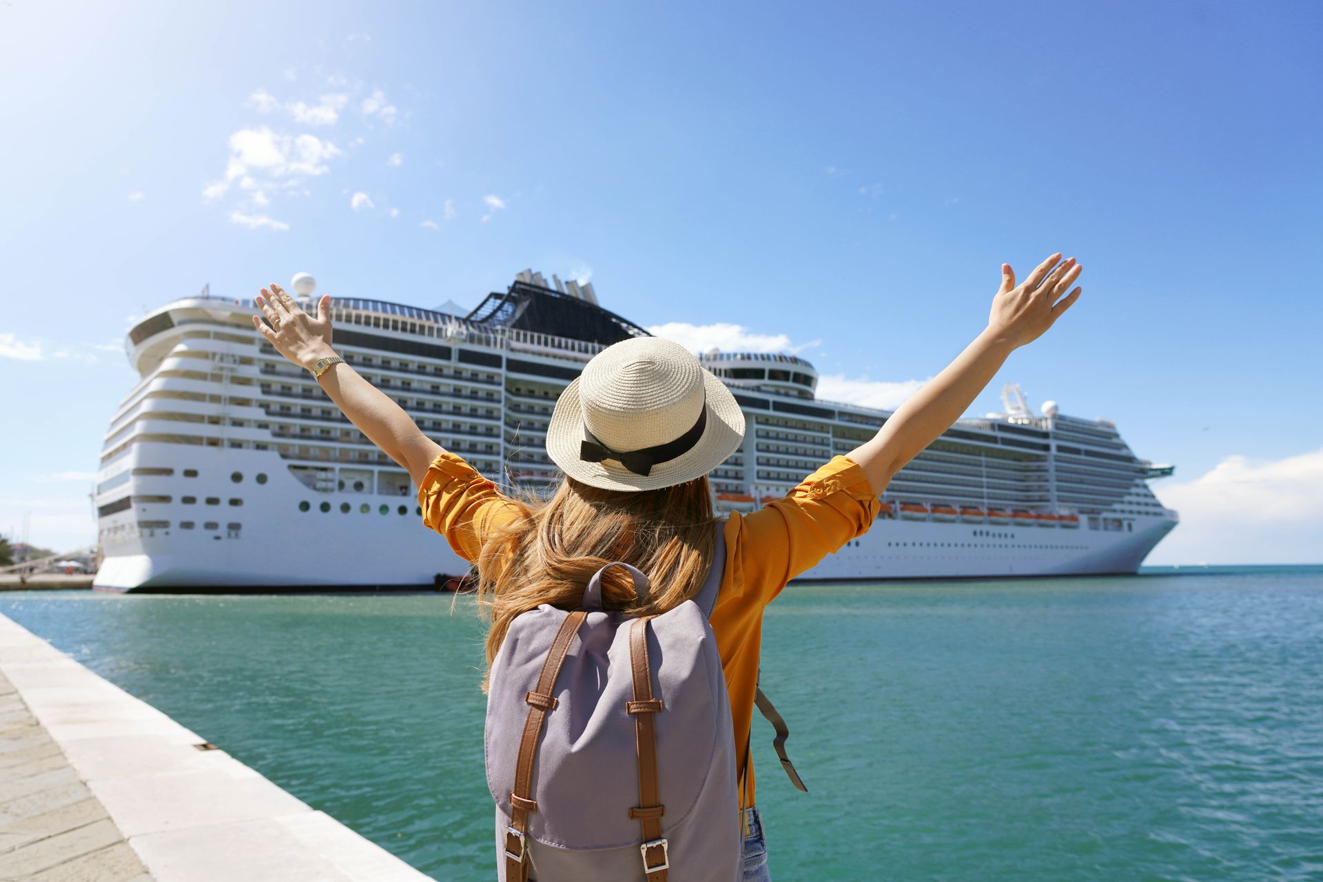 Traveler with arms raised faces a large cruise ship in a sunny harbor