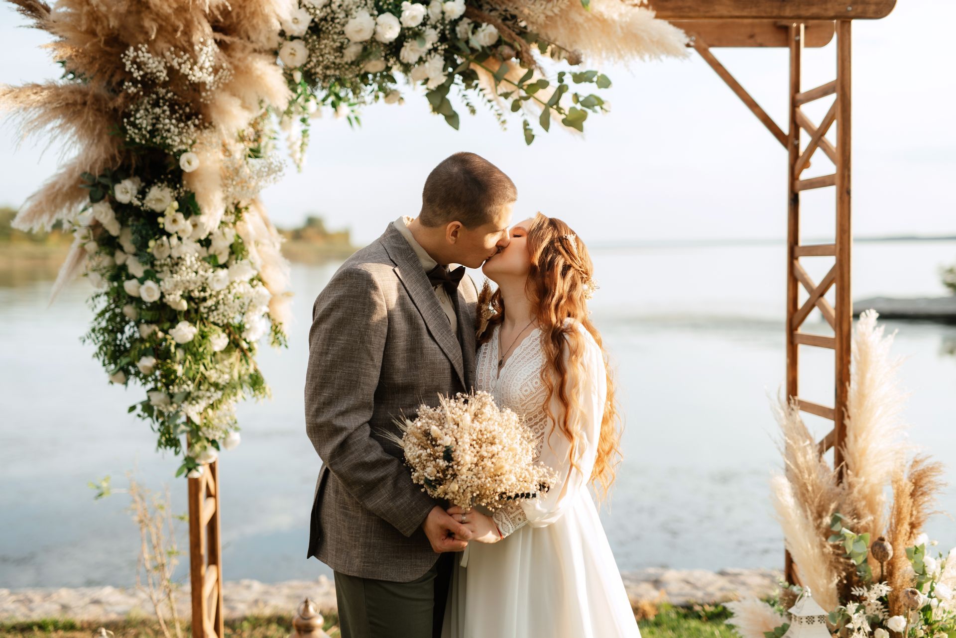Couple in wedding attire sharing a kiss by a floral arch overlooking the water