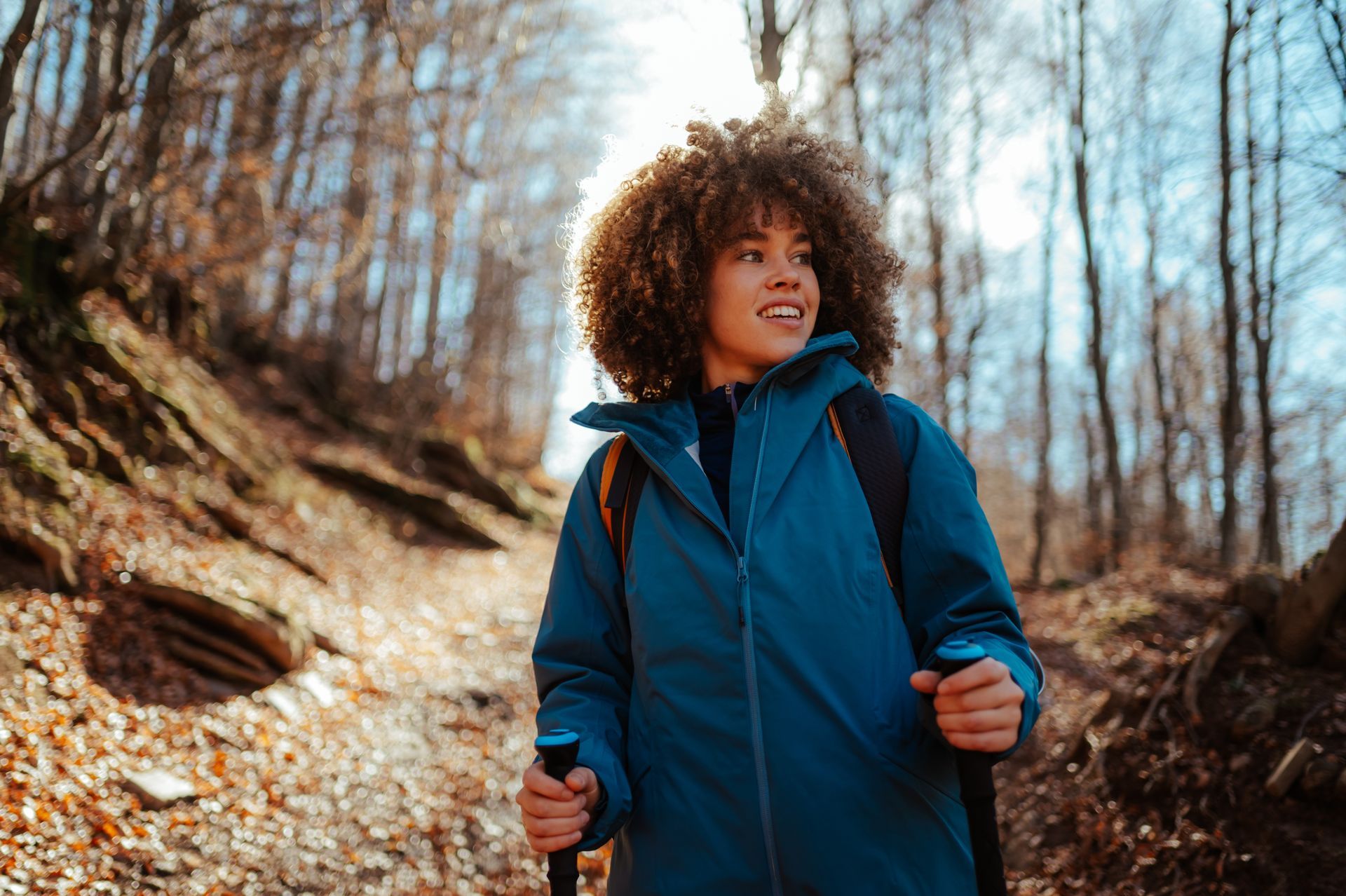 Hiker in a blue jacket walking on a wooded trail, holding trekking poles and smiling upward