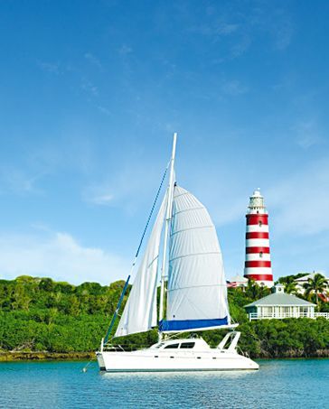 White sailboat on turquoise water with a red-and-white lighthouse on a green shore under a blue sky