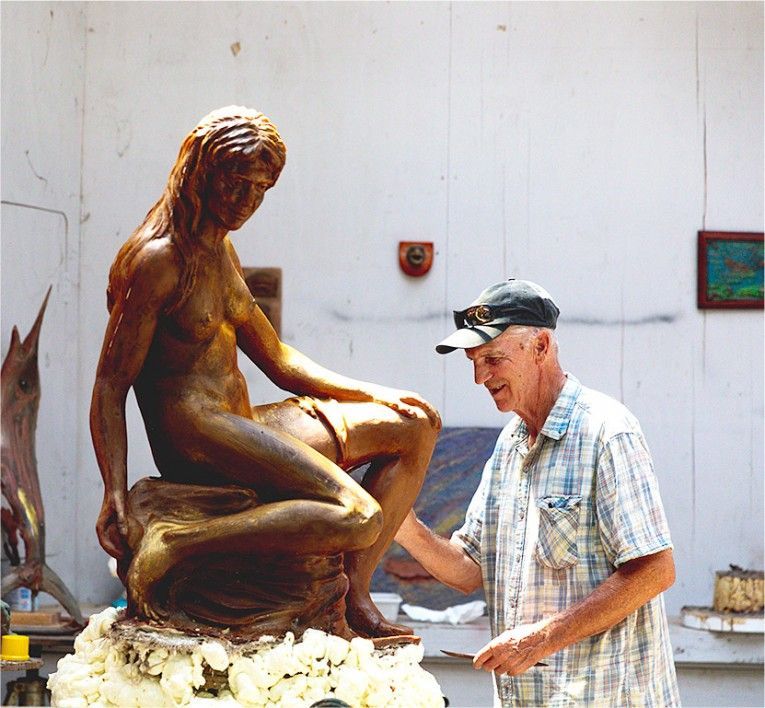 Man in cap sculpting a bronze seated figure in a workshop-like outdoor setting