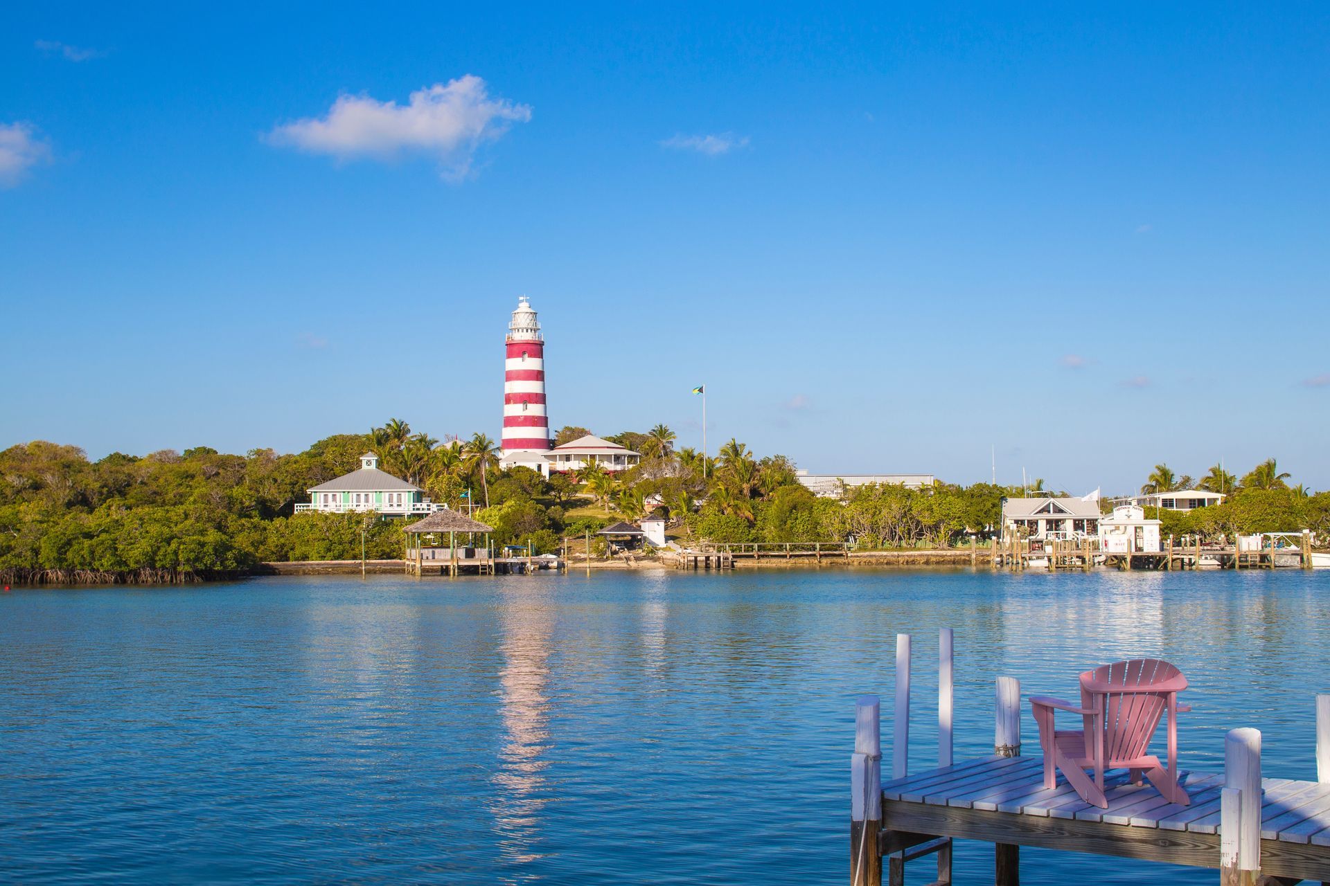 Red-and-white striped lighthouse by calm blue water with a dock in the foreground and trees behind