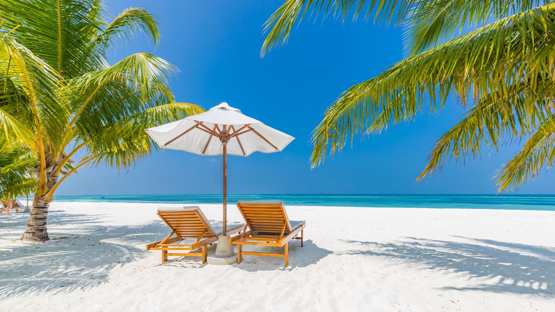 Beach scene with two lounge chairs and a white umbrella on bright white sand, framed by palm trees and turquoise water