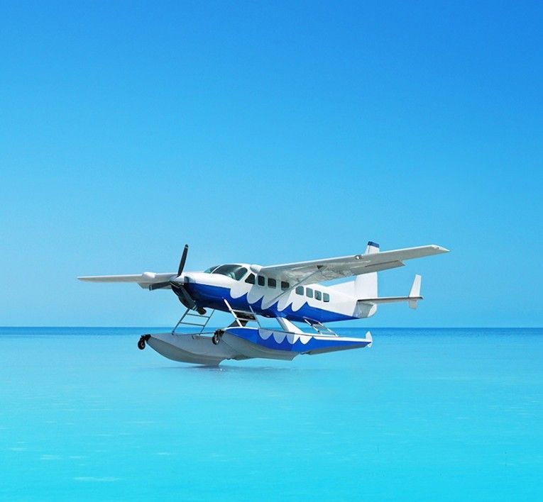 A blue-and-white seaplane flying low over calm turquoise water under a clear blue sky