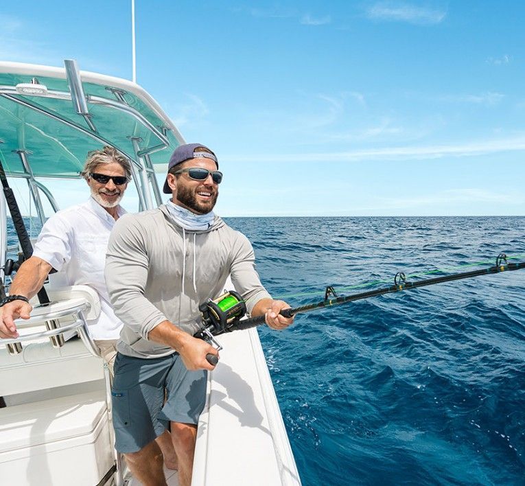 Two anglers on a boat, one reeling in a fishing rod on a bright blue ocean day.