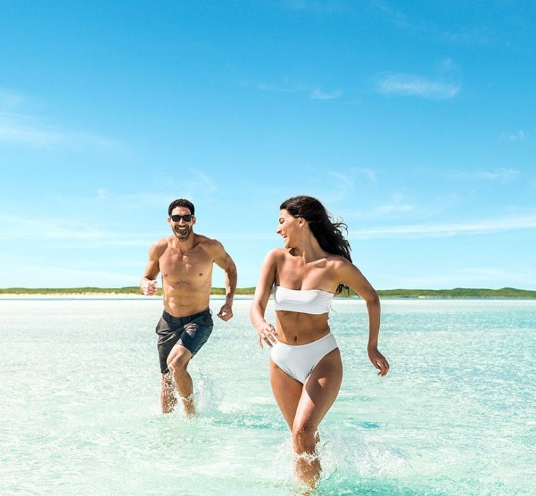 Two people running through shallow turquoise water under a bright blue sky