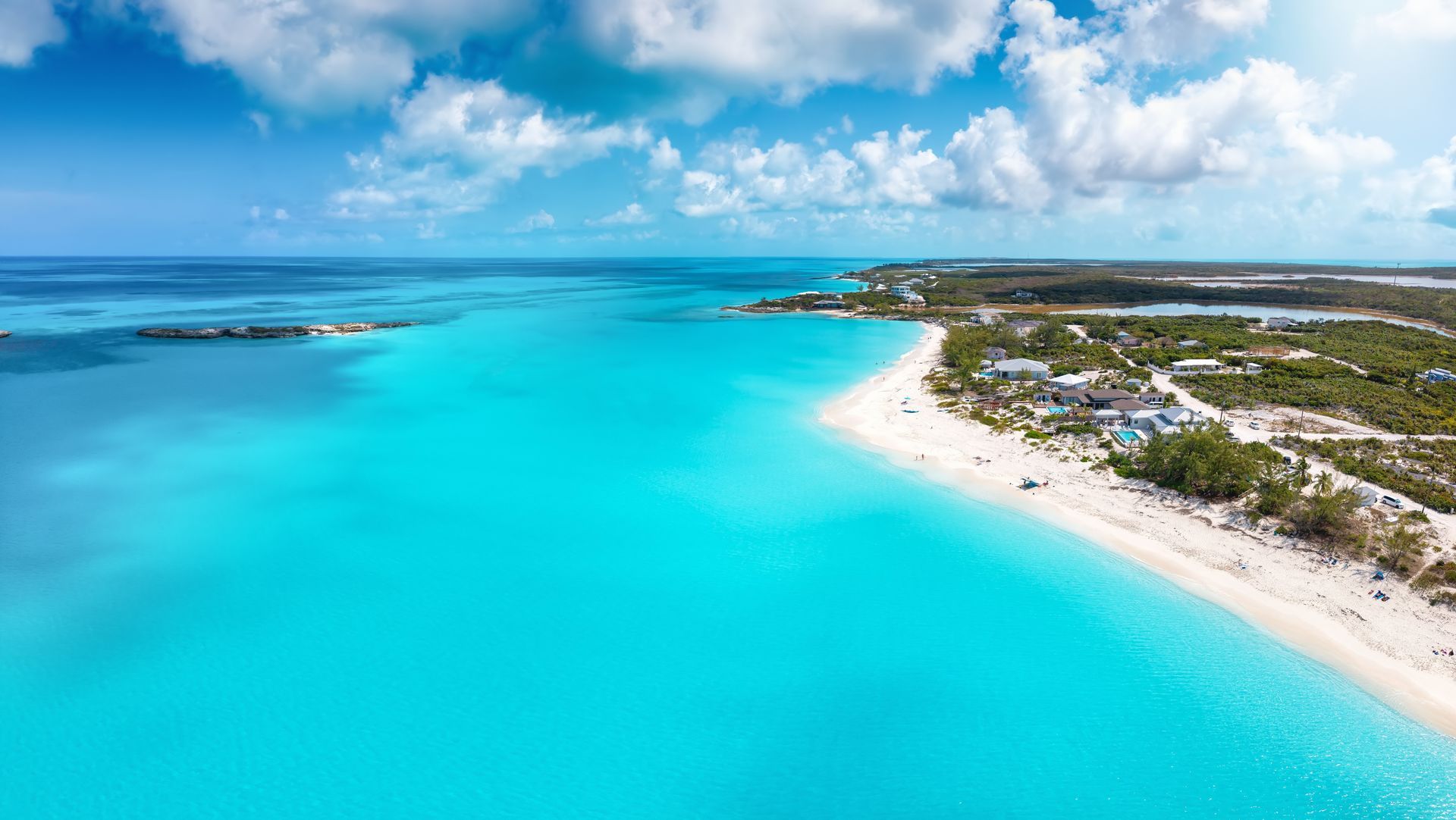 Aerial view of a turquoise tropical beach with white sand, scattered homes, and a calm blue sea