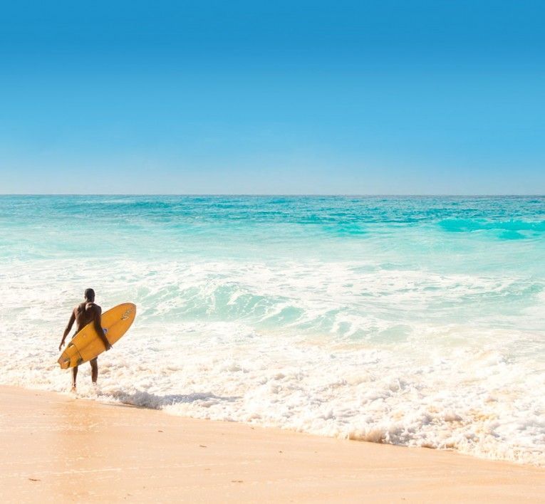 Surfer carrying a yellow surfboard along a sunny beach with turquoise waves and blue sky
