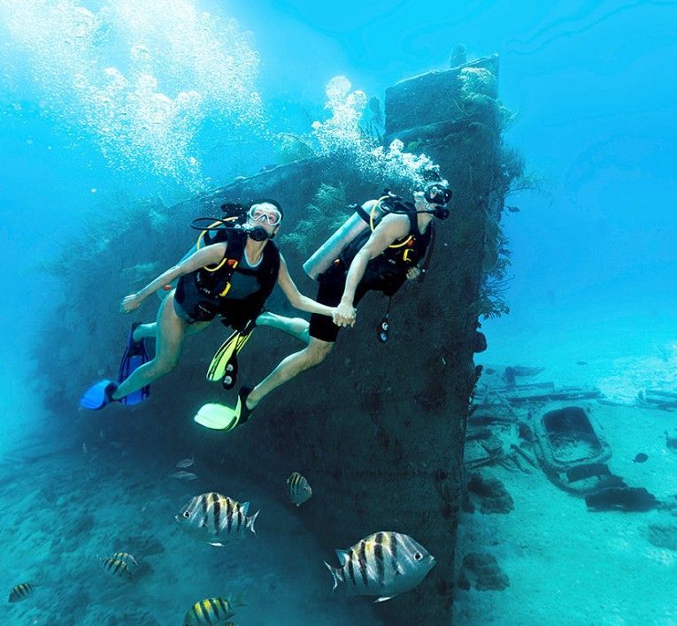 Two scuba divers swim beside a sunken shipwreck in clear blue water with fish nearby