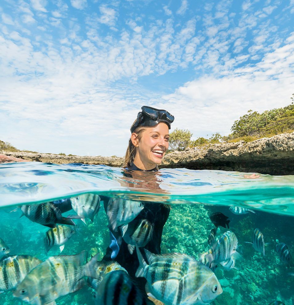 Snorkeler smiling above clear turquoise water with striped fish below and rocky shoreline under a blue sky
