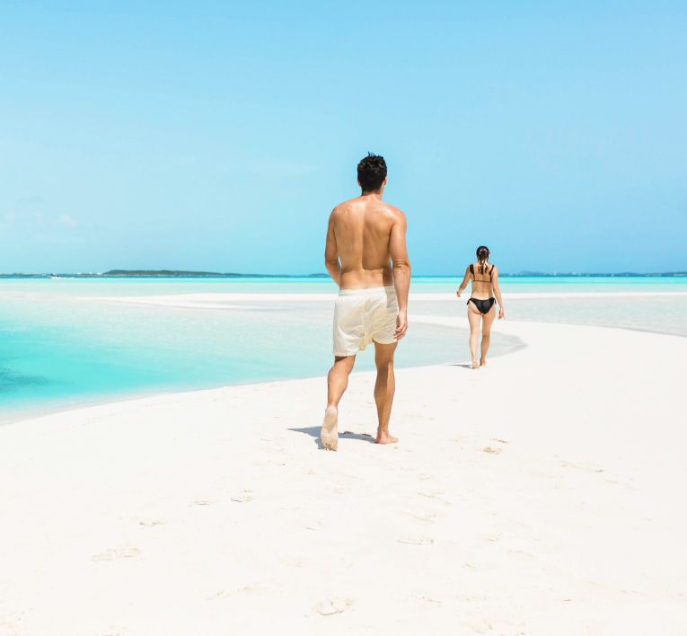 Two people walking on a white sandbar beside turquoise water under a clear blue sky