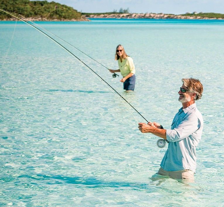 Two people fly fishing in shallow turquoise water near a tropical shoreline