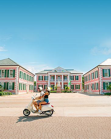 Two people ride a scooter in front of pink-and-green buildings under a clear blue sky.