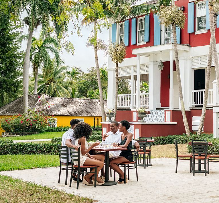 People seated at an outdoor table beside a red-and-white building in a tropical garden.