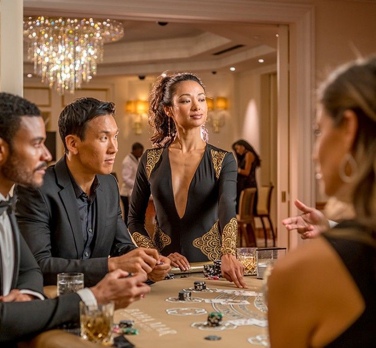 People at a casino table, with a woman in a black dress standing and speaking in a warmly lit room.