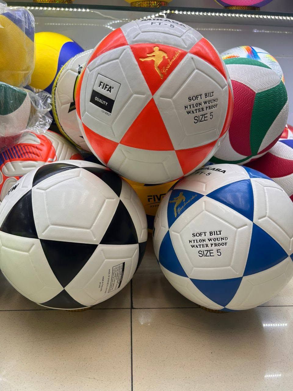 Varios balones de fútbol de colores apilados sobre un suelo de baldosas.