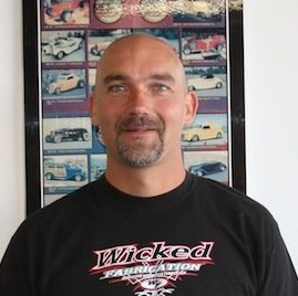 A bald man with a beard is wearing a black wicked fabrication t-shirt.