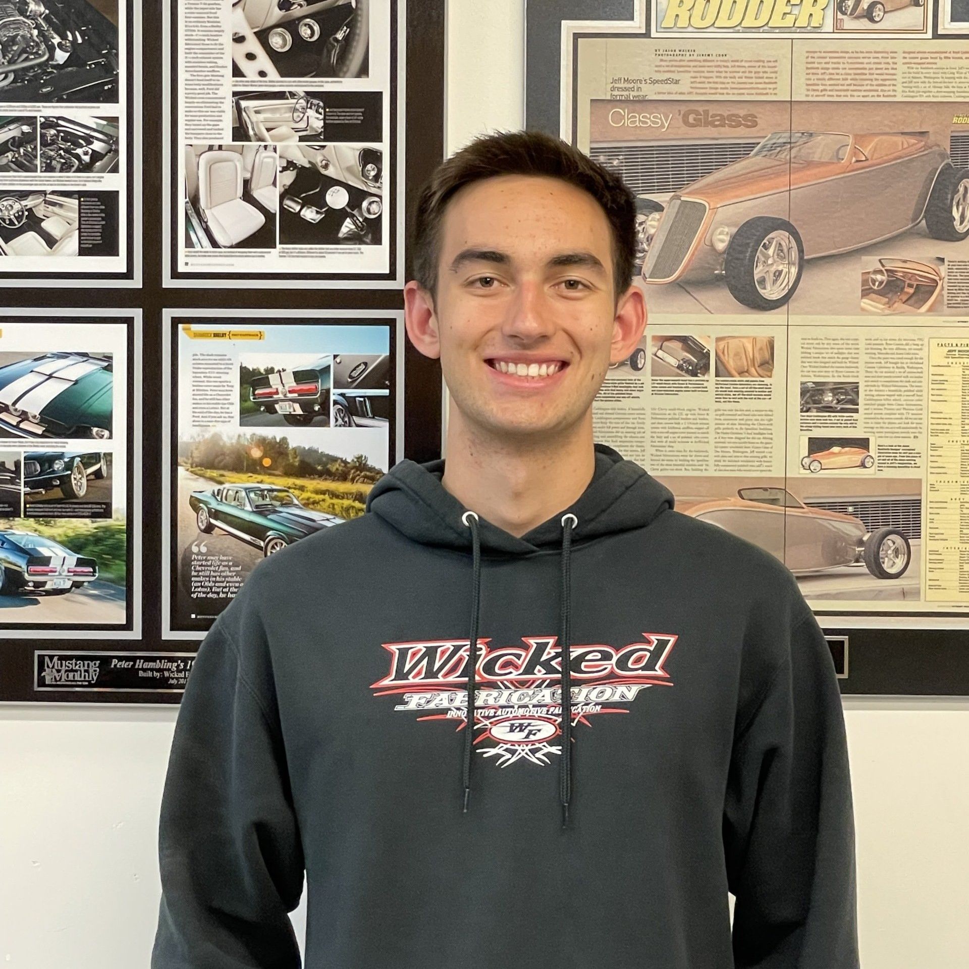 A young man wearing a hoodie is standing in front of a wall with pictures of cars on it.