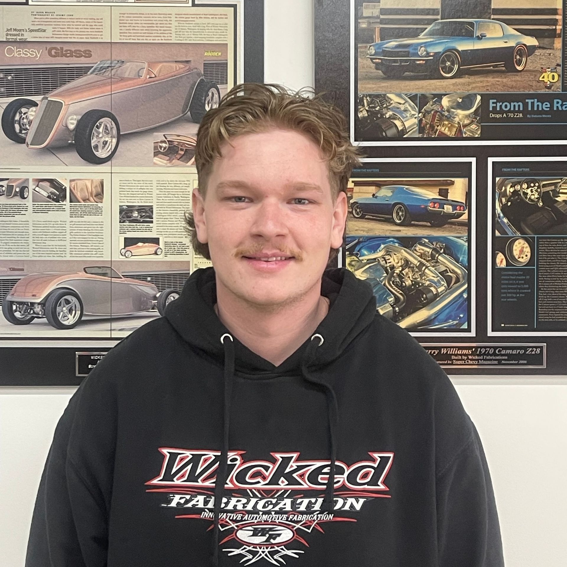 A man wearing a black hoodie is standing in front of a wall with pictures of cars on it.